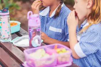 Two young girls in school uniform sit together on a bench eating packed lunches